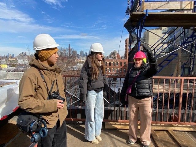 Three people wearing hard hats on the roof deck of a construction site with the New York City skyline in the background
