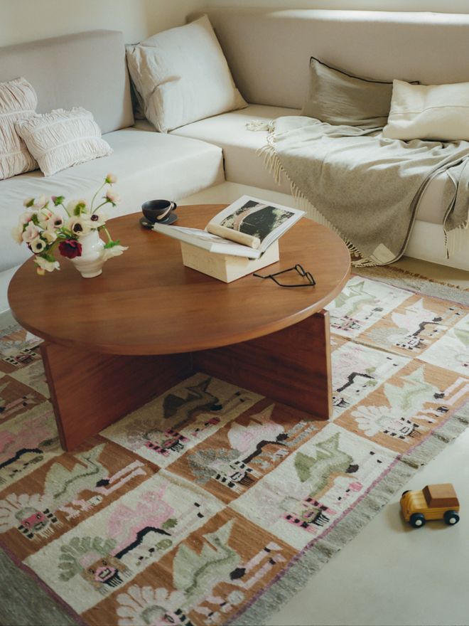 A living room corner with a light sofa layered with cushions, a round wooden coffee table with flowers and books, and a colourful patchwork-style patterned rug. A small wooden toy car sits on the floor.