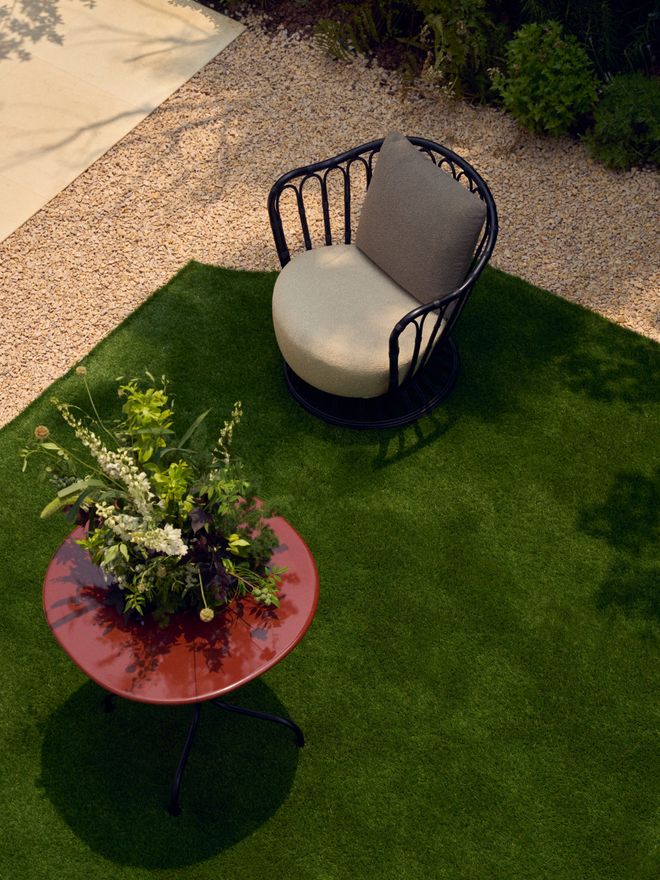 Overhead view of an outdoor lounge chair standing on a lawn beside a gravel path. In front of it, a round red side table holds an arrangement of white flowers and greenery.