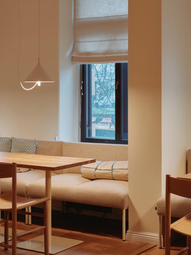 Valoisa ja lämmintunnelmainen working café -nurkkaus, jossa sisustuksena puinen pöytä ja tuoli, pitkä pehmustettu penkki sekä katossa kartiomainen riippuvalaisin.