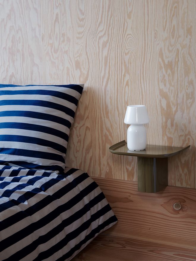 HAY's Korpus shelf is attached to the bedroom wall next to the bed as a bedside table. On top of the green shelf is a white Apollo table lamp.
