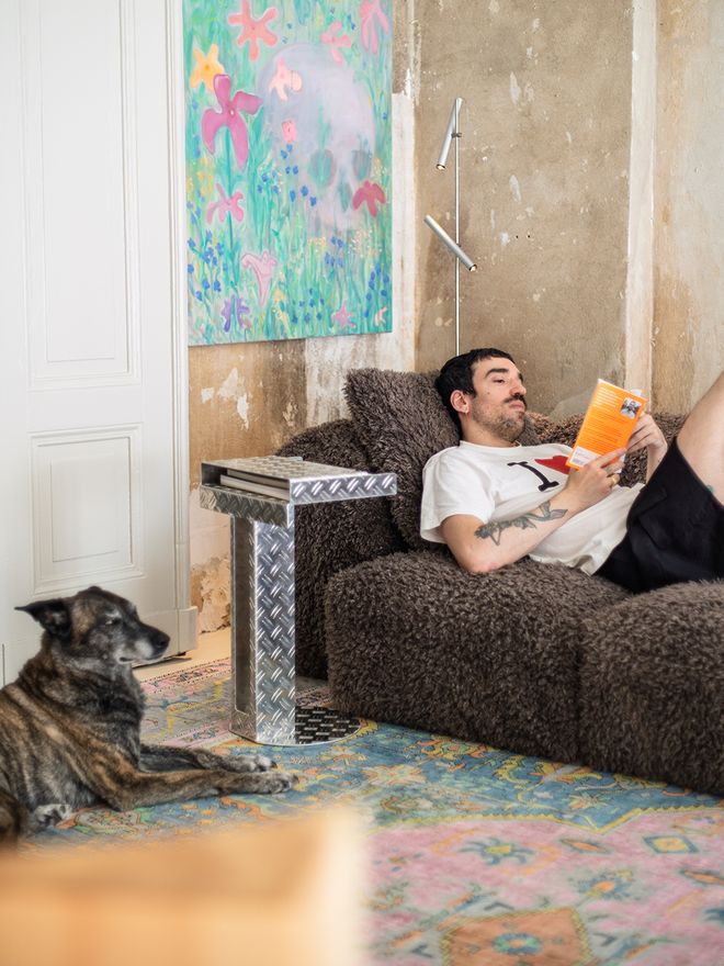 Guy reading on a brown sofa with Frecha the dog lying on a patterned rug in the foreground