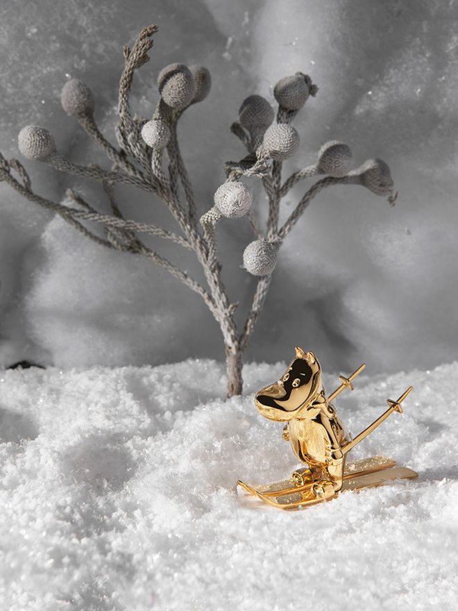A gilded Moomintroll figurine with skis and sticks in a wintery landscape.