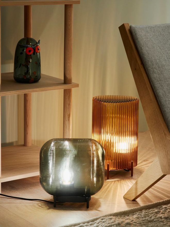Copper-toned Putki table lamp and a gray glass lamp on the floor beside a wooden shelf and a lounge chair. A glass owl sits on the shelf.