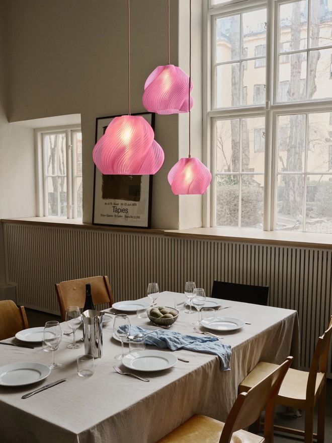 Three pink Soft Serve pendans hanging above a dining table, casting a soft glow in a bright room framed by large windows
