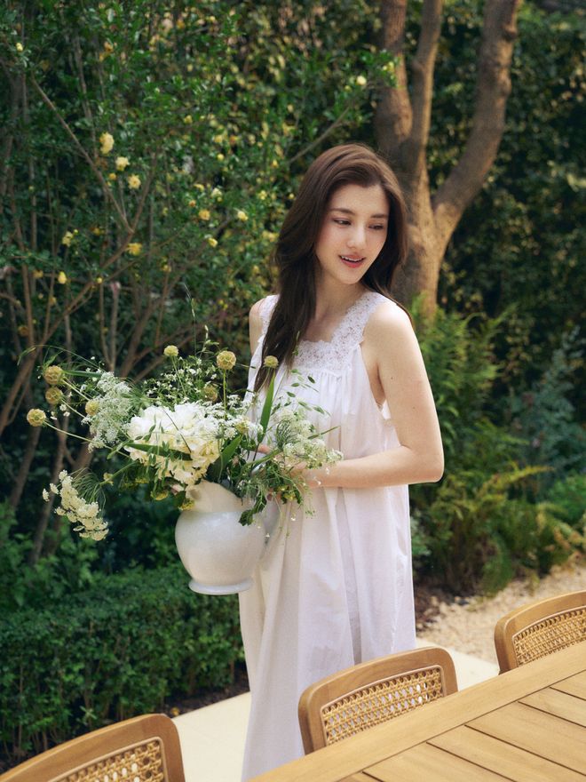 Karena Ng a white sleeveless dress carrying a vase of flowers past a wooden dining table with woven chairs, with trees behind her.