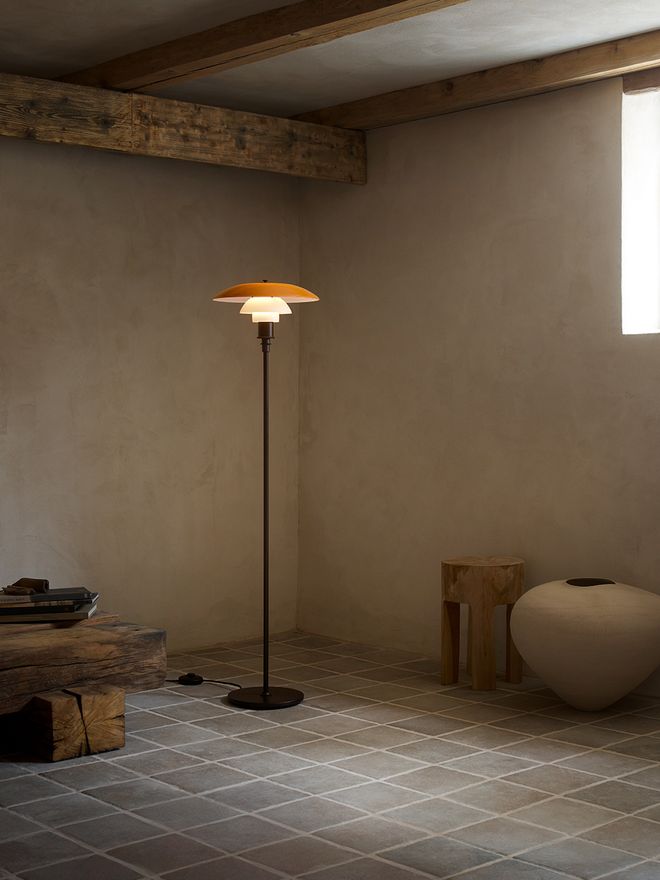 Louis Poulsen PH 3½-2½ Floor Lamp Centenary Edition in aged brass, with tiered amber and white opal shades, standing in a minimalist interior with exposed wooden ceiling beams and diagonal tile flooring