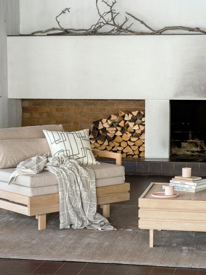 A cushion and a blanket on a Day&Night chair bed and books and coffee cups on a wooden Day&Night coffee table. In the background, a fireplace and a pile of firewood.