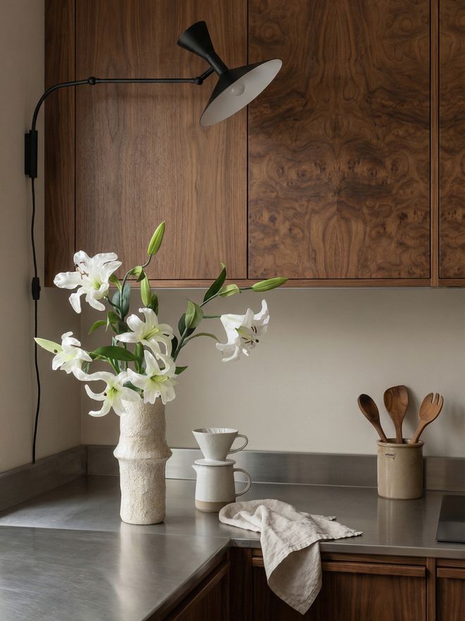 A bouquet of white lilies in a pale ceramic vase on a kitchen counter, with dark wooden cabinets and a black wall lamp in the background.