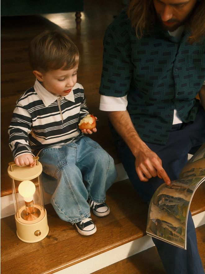 A child sitting on the stairs next to a yellow The Muse lamp while an adult shows them a book.