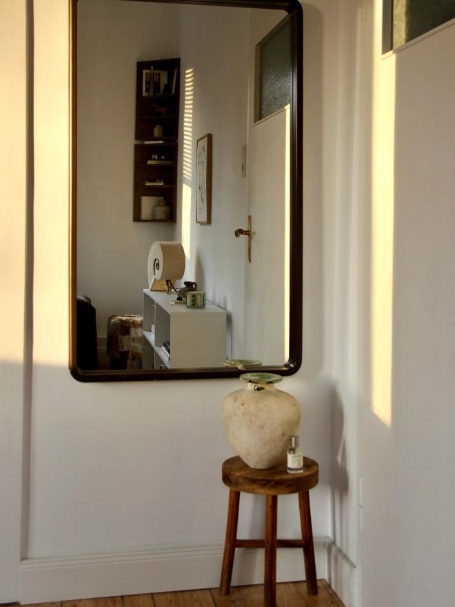 An entryway corner with a large dark-framed wall mirror above a wooden stool holding a pale ceramic vase.