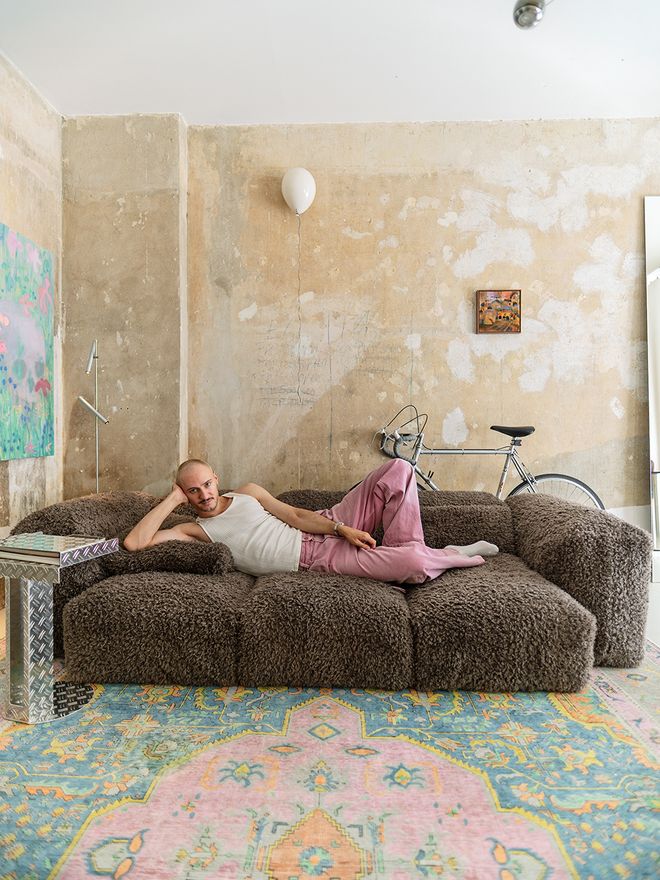 Dima lying on the gray sofa in the living room, with a bicycle leaning against the raw plaster wall behind him
