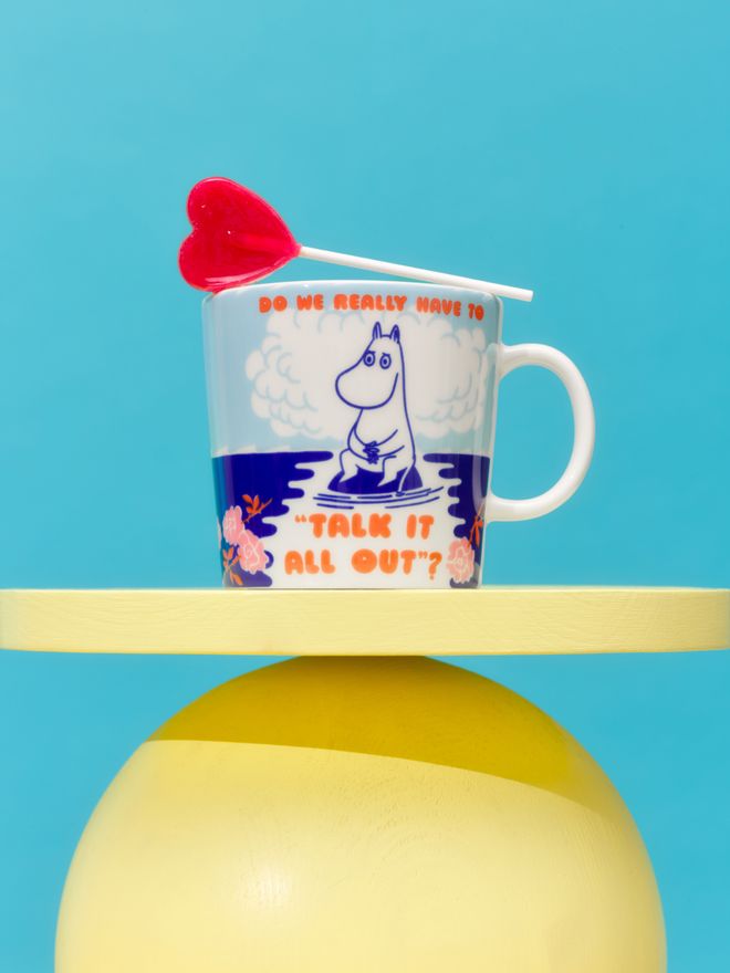 A patterned “Talk it all Out” Moomin mug on a yellow base against a blue background, with a red heart-shaped lollipop leaning against the mug.