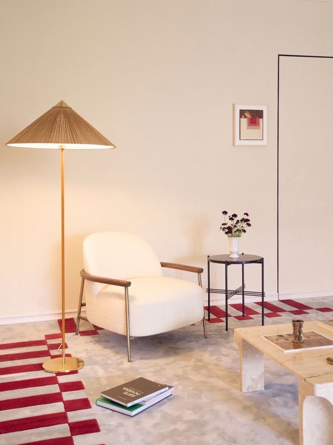 White lounge chair beside a brass floor lamp with a cone-shaped shade and a small side table with flowers, on a red and white patterned rug.
