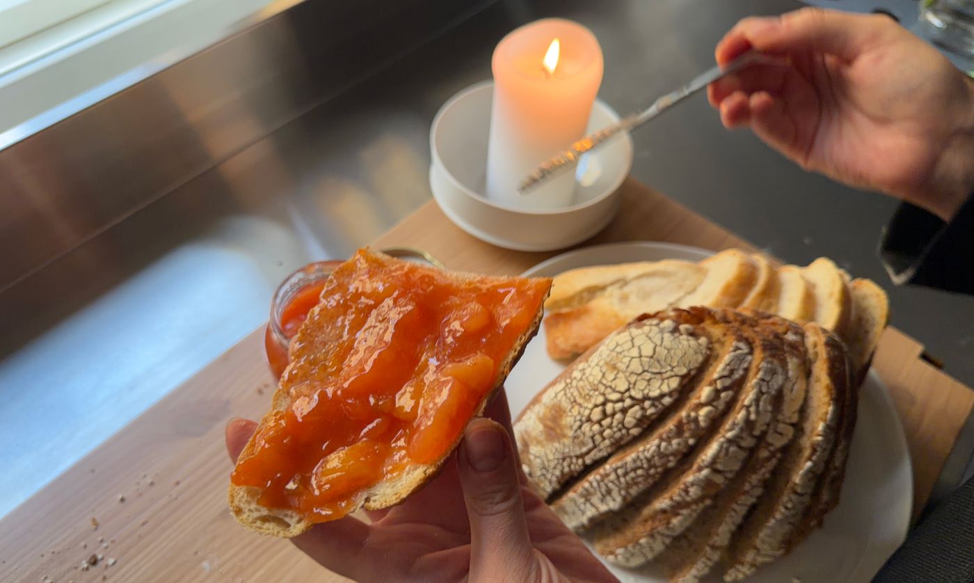 Käsi pitelee luumuhillolla päällystettyä leipäviipaletta, taustalla leipää ja kynttilä