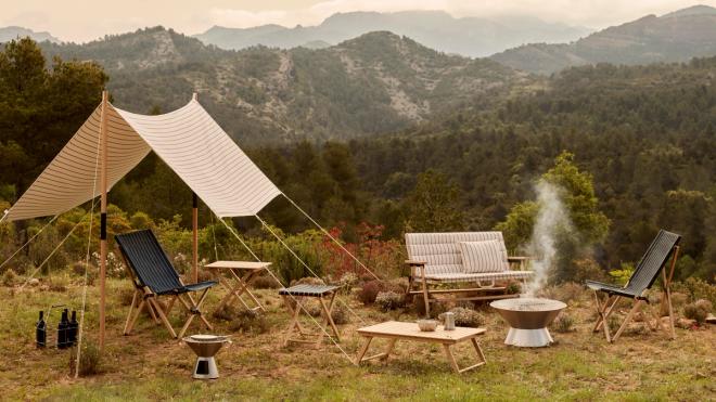 A mountain landscape featuring furniture from HAY’s Outdoor Market collection: sun loungers, folding tables, an awning, and a low fire pit with smoke rising from it.