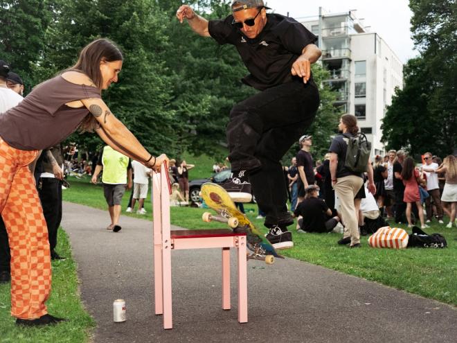 A skateboarder jumps over a pink chair held steady by another person