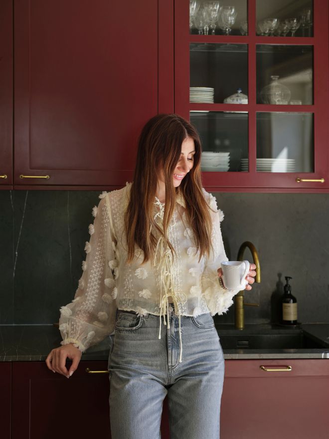 Nadia holding a coffee cup in front of deep red kitchen cabinets.