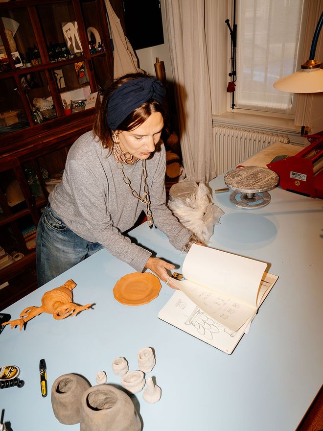 Marie is leafing through a sketchbook at her worktable, clay pieces and tools spread across it.