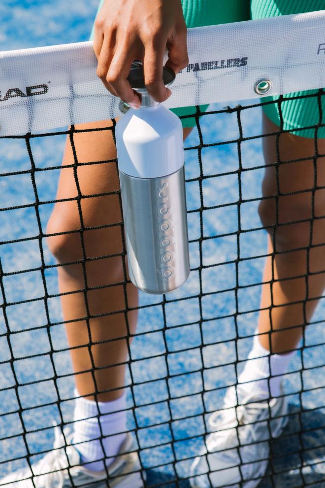 A tennis player behind the net holding a silver water bottle and a racket.