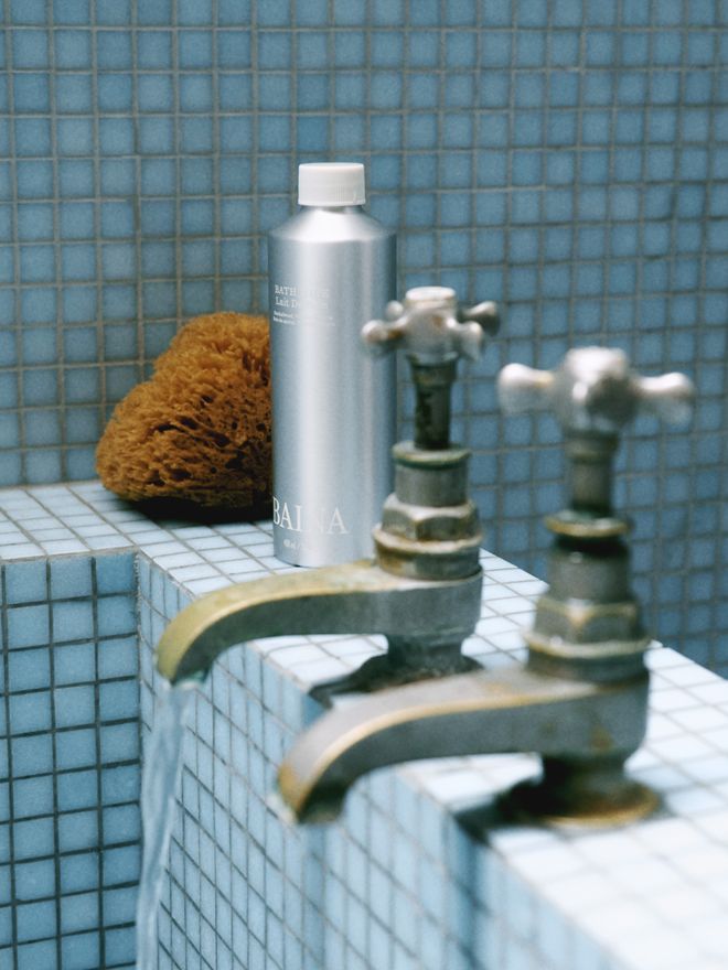 An aluminium skincare bottle and a natural sponge beside vintage brass taps, against a blue mosaic-tiled background