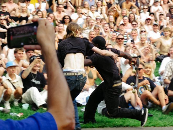 Two skateboarders cross the finish line together, side by side, as the crowd cheers on the background