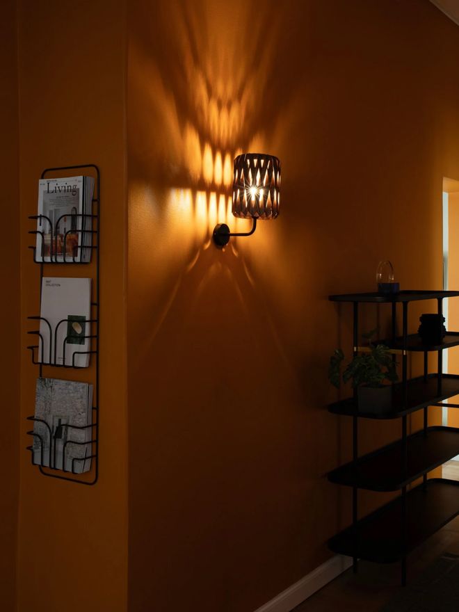 Pilke Signature wall lamp casting light patterns onto an ochre wall, next to a black magazine rack and a shelf.