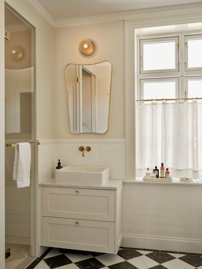 A light bathroom with a black-and-white chequered floor, an organically shaped brass-framed mirror, and a small glass light above it.