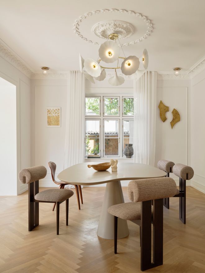 A light-filled dining room with an organically shaped table, upholstered beige chairs, and a branching chandelier with multiple globe lights overhead.