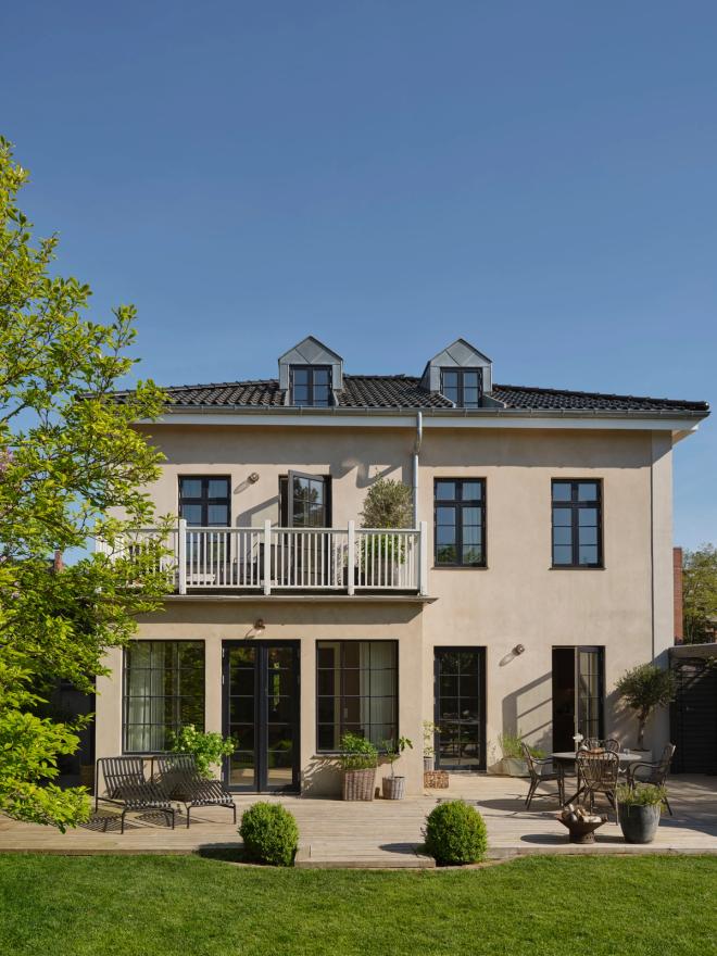 A two-storey villa in a summery garden setting, with outdoor furniture on the terrace.