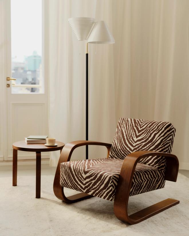 The Tank armchair with a dark wooden frame upholstered in Zebra fabric, next to a low round side table with books and a coffee cup, and a floor lamp behind it.
