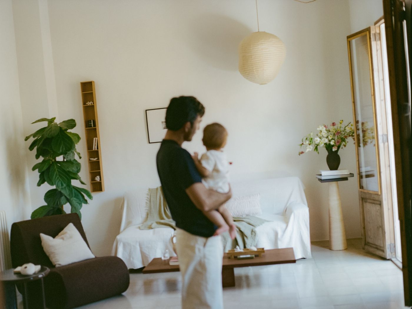Mateo stands in the living room holding a child. A pale sofa, a large houseplant, a round paper lantern overhead complete the scene.