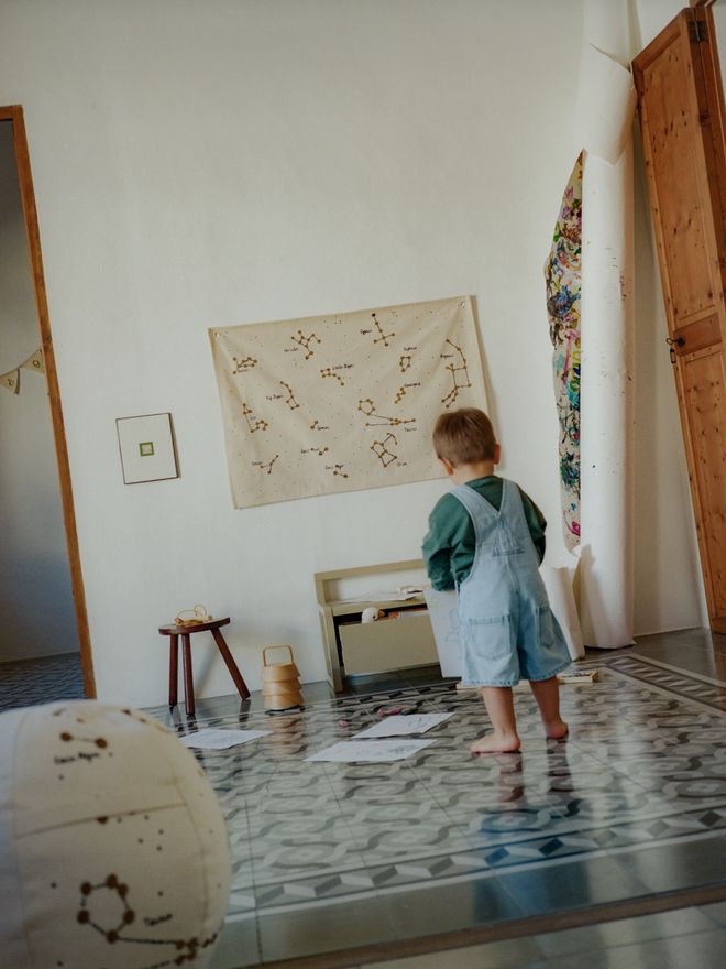 A child stands on a patterned tile floor in the nursery. A wooden stool, a wall textile, and a large ball are in the background.