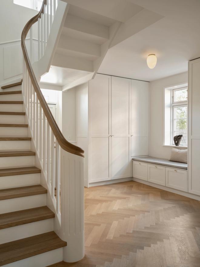 A light-filled entrance hall with a curved wooden staircase, white built-in cabinets, herringbone parquet flooring, and a small round pendant light overhead.