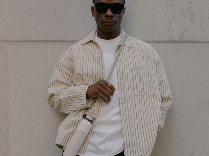 A person leaning against a light wall in a striped shirt and sunglasses, carrying a silver water bottle on a cord over their shoulder.