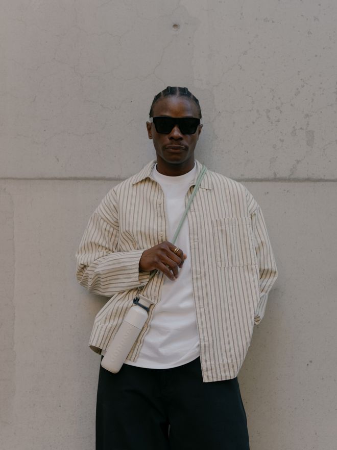 A person leaning against a light-toned wall, wearing a striped shirt and sunglasses, with a water bottle hanging from a cord over their shoulder.
