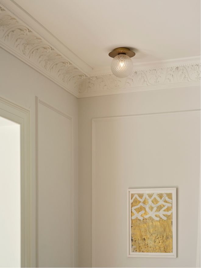 The upper corner of a room featuring a decorative ceiling moulding, a small round glass ceiling light, and a golden patterned artwork on the wall.