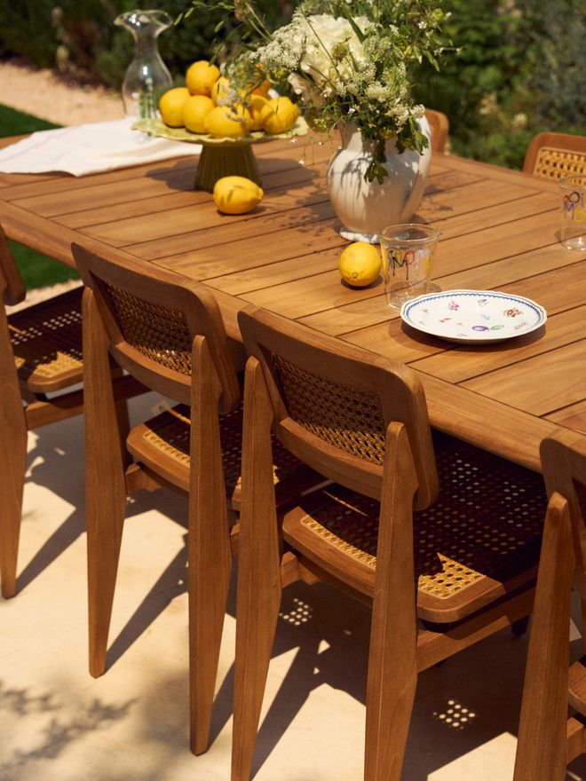 A wooden dining table set with lemons, a vase of white flowers, and a floral plate, surrounded by wooden chairs with woven seats and backs, bathed in sunlight.