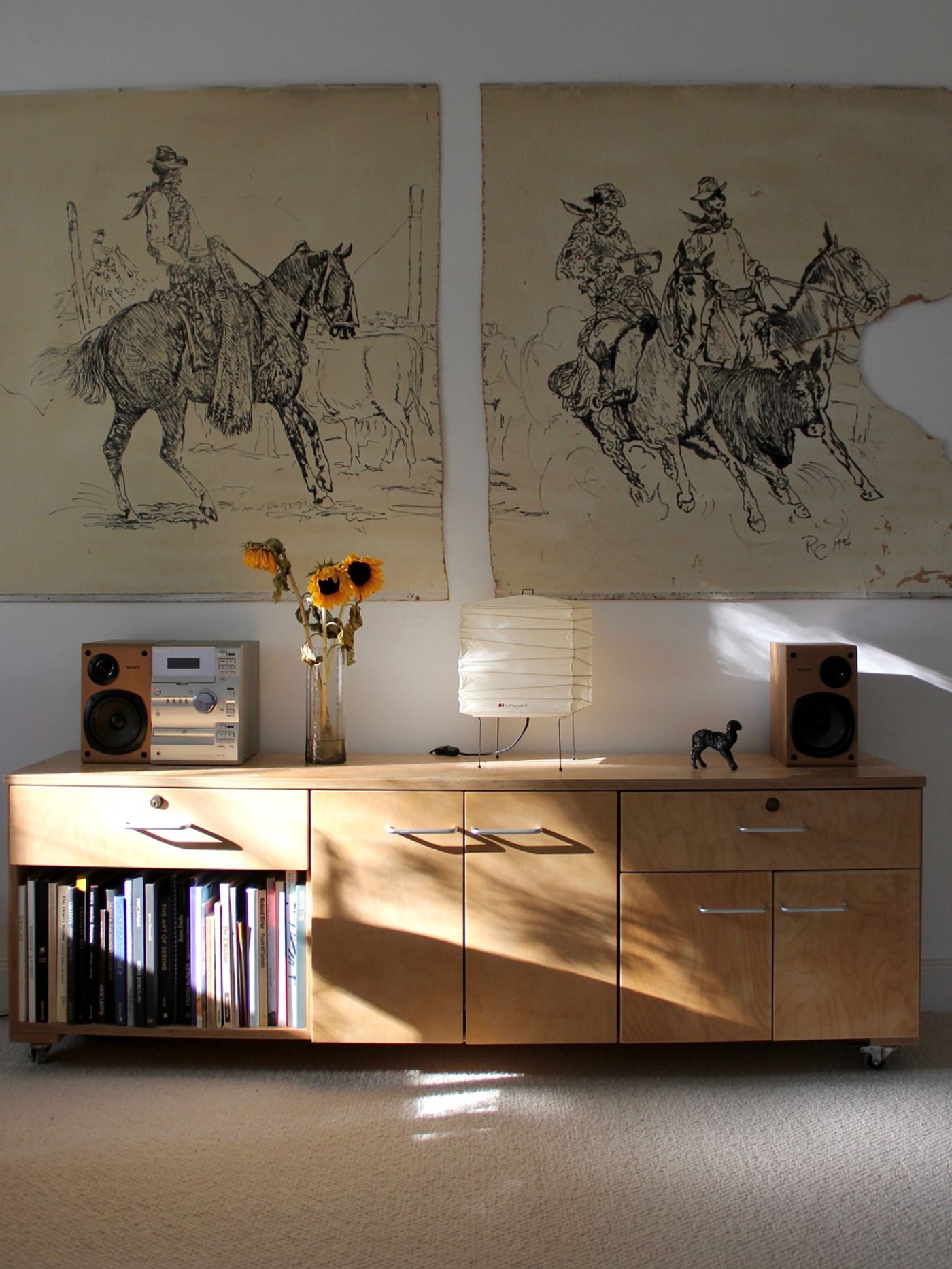 A wooden sideboard with a stereo, speakers, and a bouquet of sunflowers, above hangs a large drawing of horses and riders.
