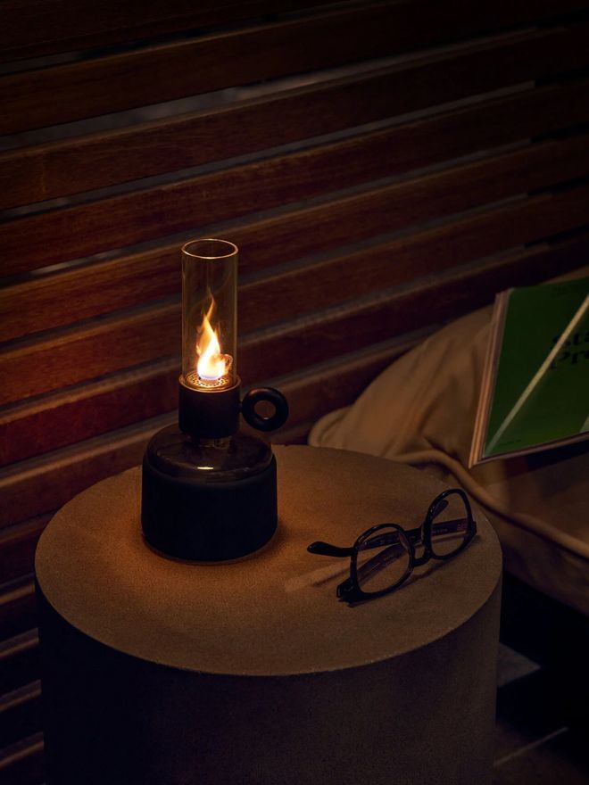 Black Flamtastique oil lamp burning on a wooden side table next to a pair of glasses in a dimly lit room.