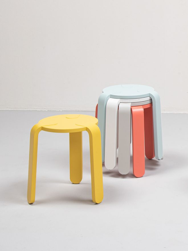A bright yellow three-legged Lulu stool stands in the foreground, with a colorful stack of similar stools in different shades forming a playful tower in the background.