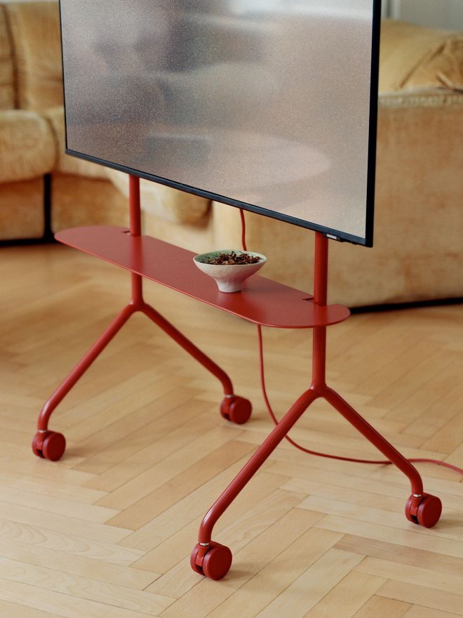 A close-up of the lower section of a red TV stand. A small bowl sits on its shelf, with a herringbone floor and a beige sofa in the background.