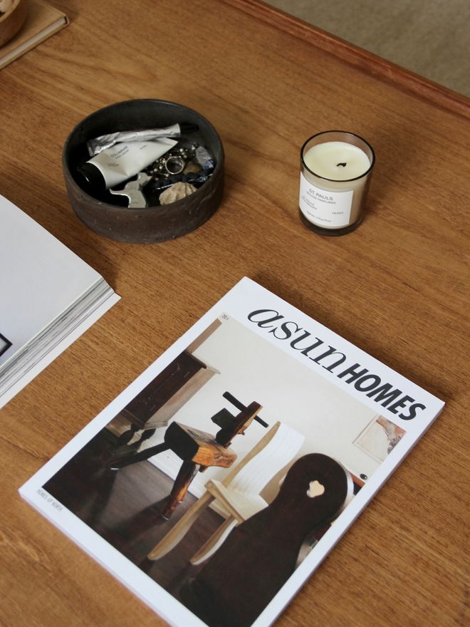 A dark ceramic bowl of jewellery, a white candle, and a stack of magazines on a wooden table.