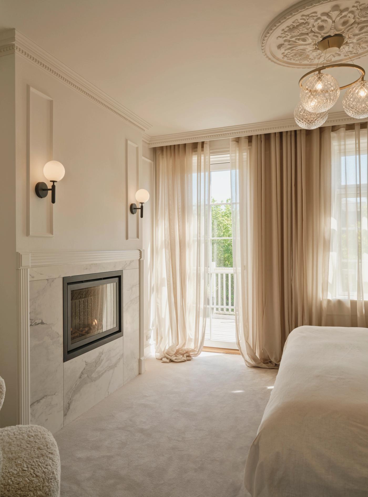 A light bedroom with a marble fireplace, globe wall lamps, and large windows.
