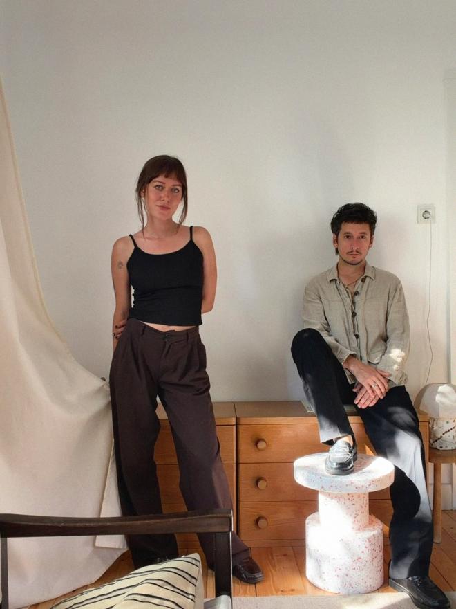 A woman standing and a man sitting on a wooden chest of drawers in a bright room.