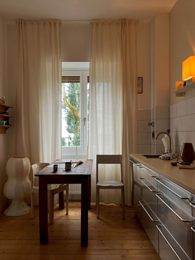 A kitchen with a dark wooden dining table by the window, pale curtains, a steel counter, and wooden floor.