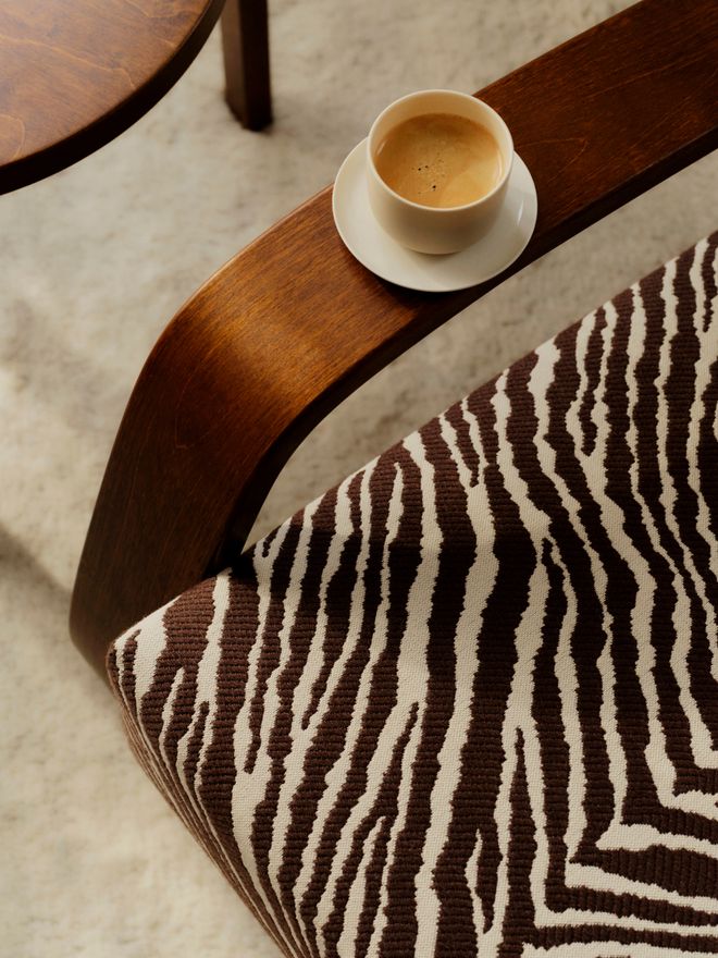 A close-up of the Tankki armchair's dark wooden armrest and Zebra-patterned seat, with a coffee cup and saucer resting on the armrest.