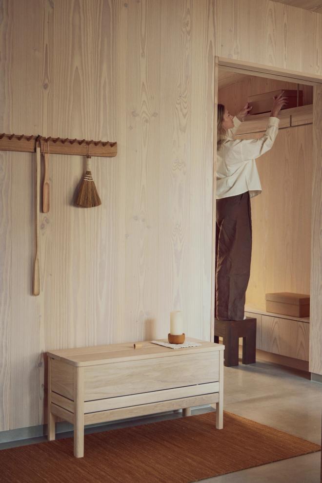 A light wooden storage bench in a pale-toned entry space, with a wall hook rail and a doorway in the background where a person is organising items.