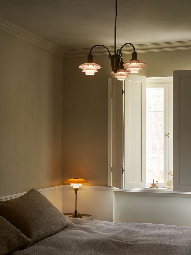 Louis Poulsen PH 1/1 Chandelier Centenary Edition with three arms in aged brass and terracotta opal glass shades, suspended above a minimalist bedroom with French doors and natural light