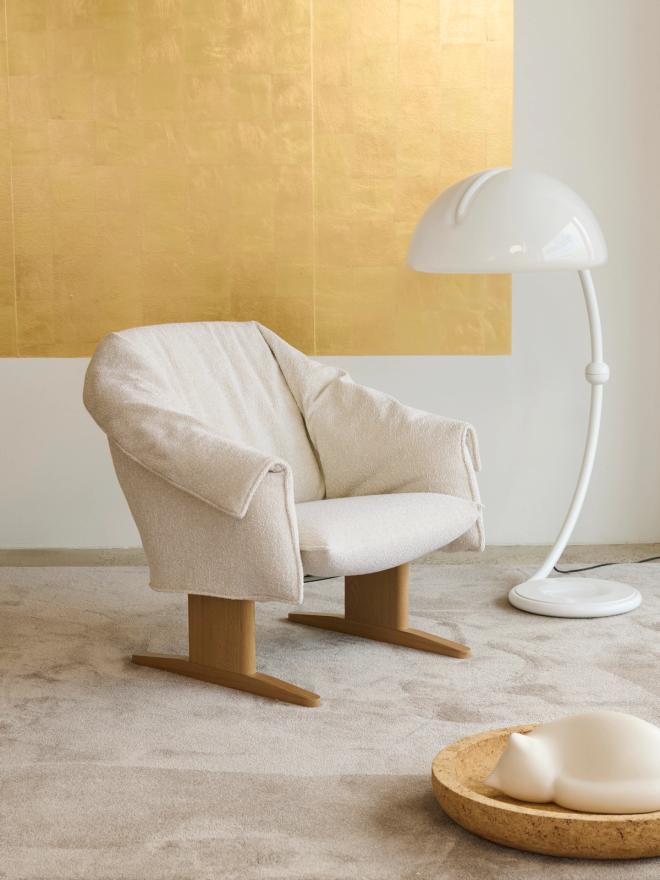 An off-white Bascule lounge chair on wooden legs in front of a gold wall, with a curved white floor lamp beside it.